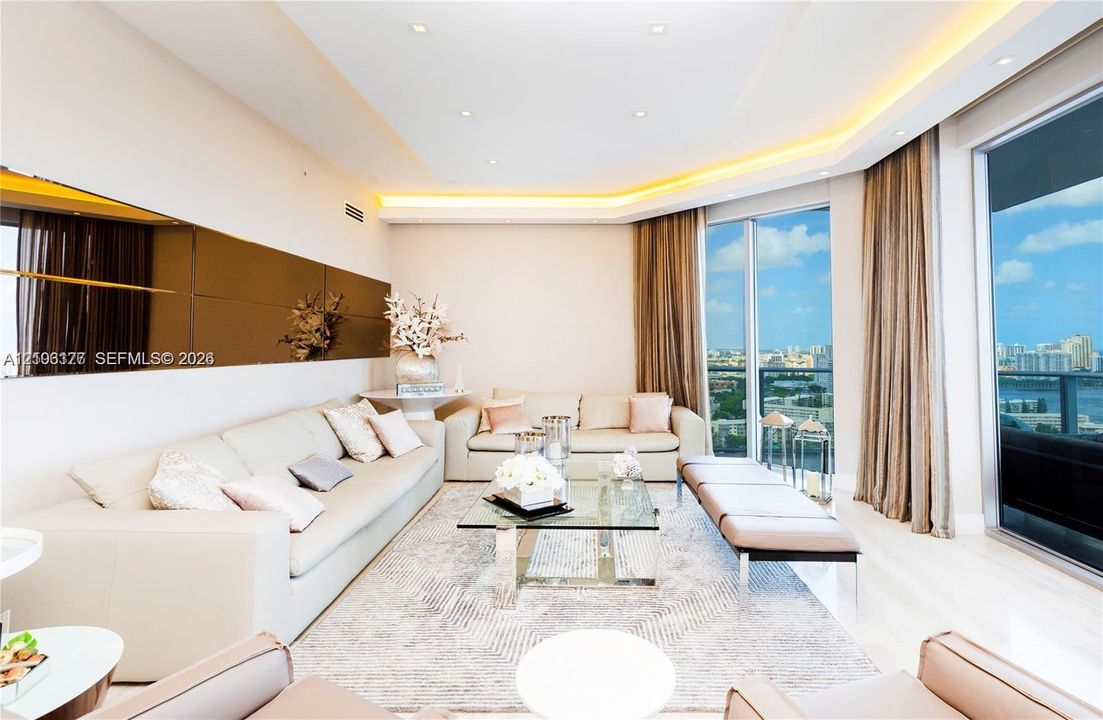 Floor-to-ceiling glass sliding doors, recessed tray ceiling with integrated cove lighting, light-toned polished flooring, mirrored wall paneling, and an expansive balcony