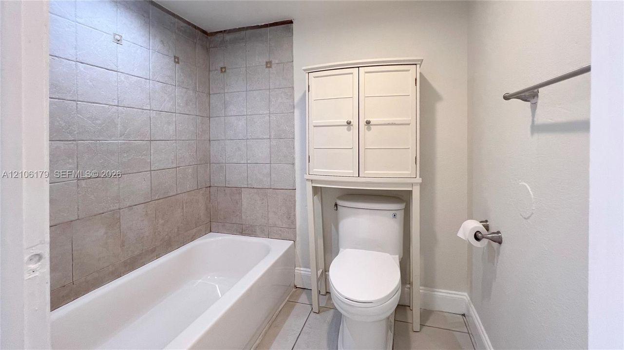 Full bathroom featuring a white porcelain tub with neutral tiled surround, over-the-toilet storage cabinet, and large-format tile flooring