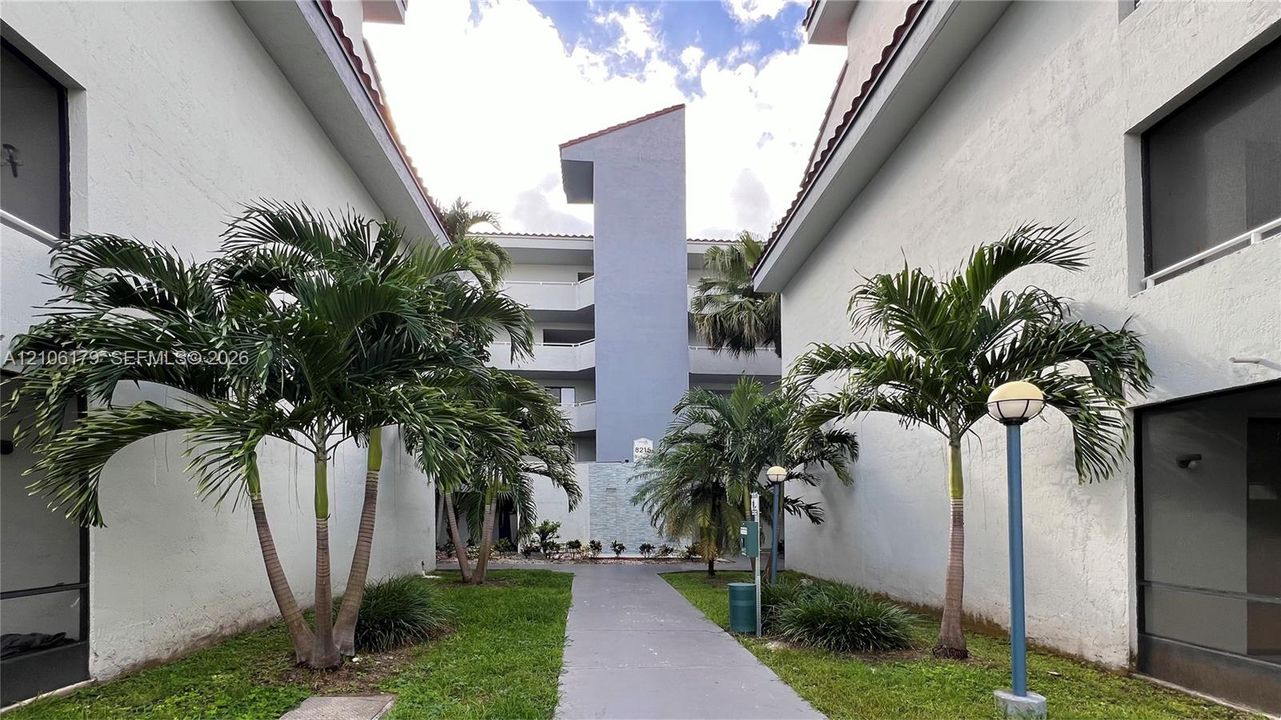 Multi-story stucco exterior featuring a paved walkway, mature palm trees, manicured landscaping, and post-mounted outdoor lighting