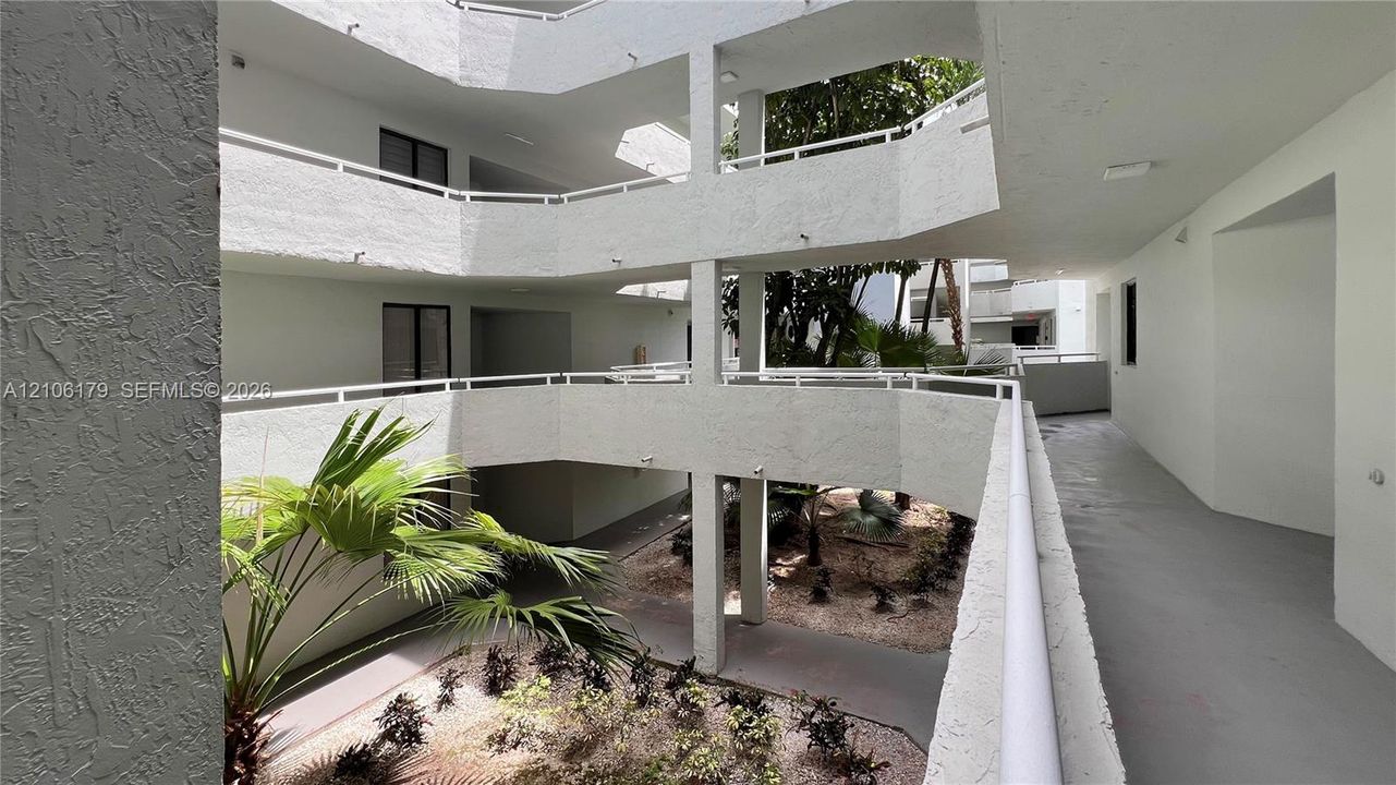 Multi-story open-air atrium featuring stucco wall finishes, metal railings, and a central landscaped courtyard with palm trees