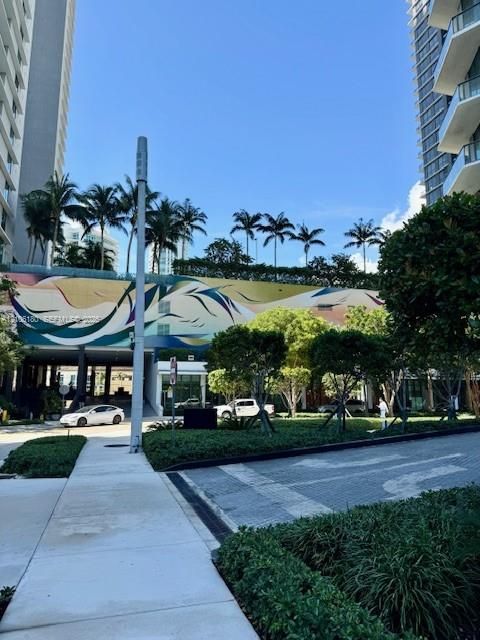 Exterior grounds featuring high-rise building facades, a decorative mural, mature palm trees, landscaped shrubbery, and paved pedestrian walkways