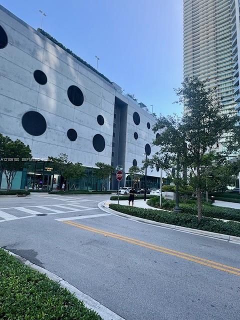 Multi-level concrete structure featuring a geometric circular facade, ground-level glass storefronts, and landscaped perimeter hedging