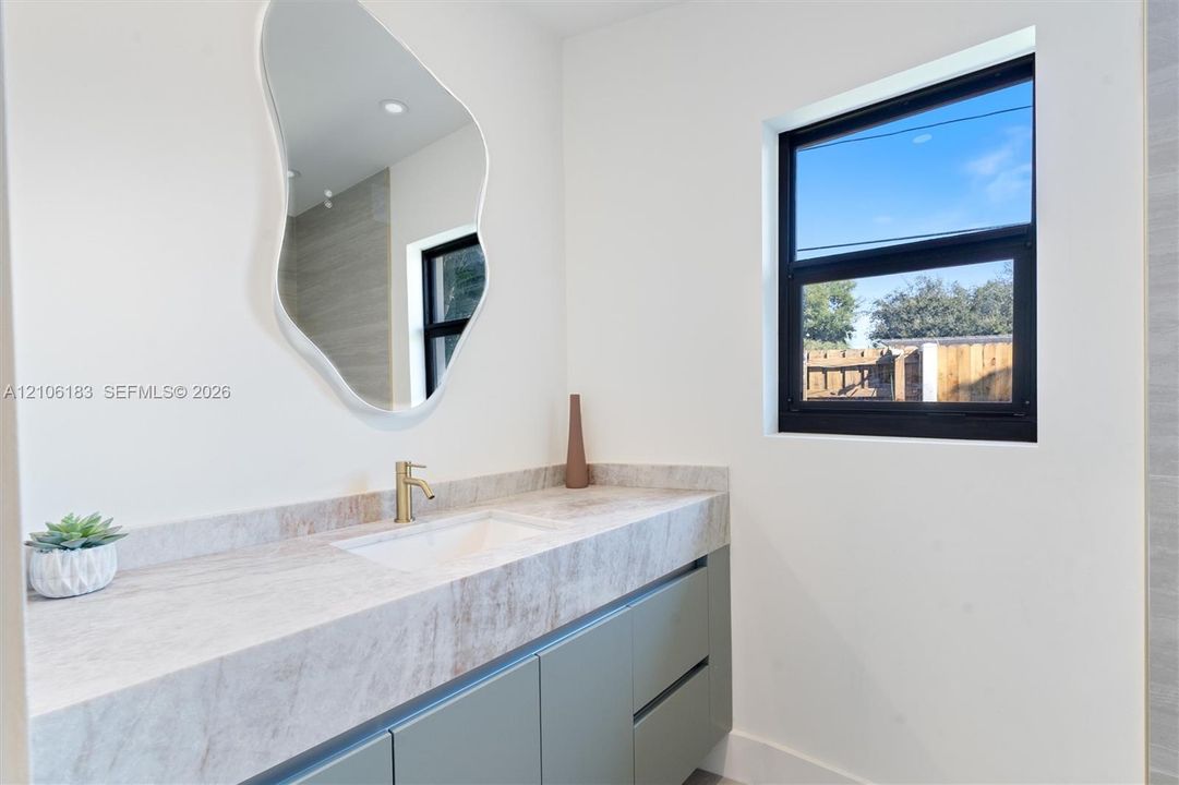 Wall-mounted vanity with solid-surface countertop, integrated rectangular sink, brass-finish faucet, organic-shaped mirror, and black-framed window