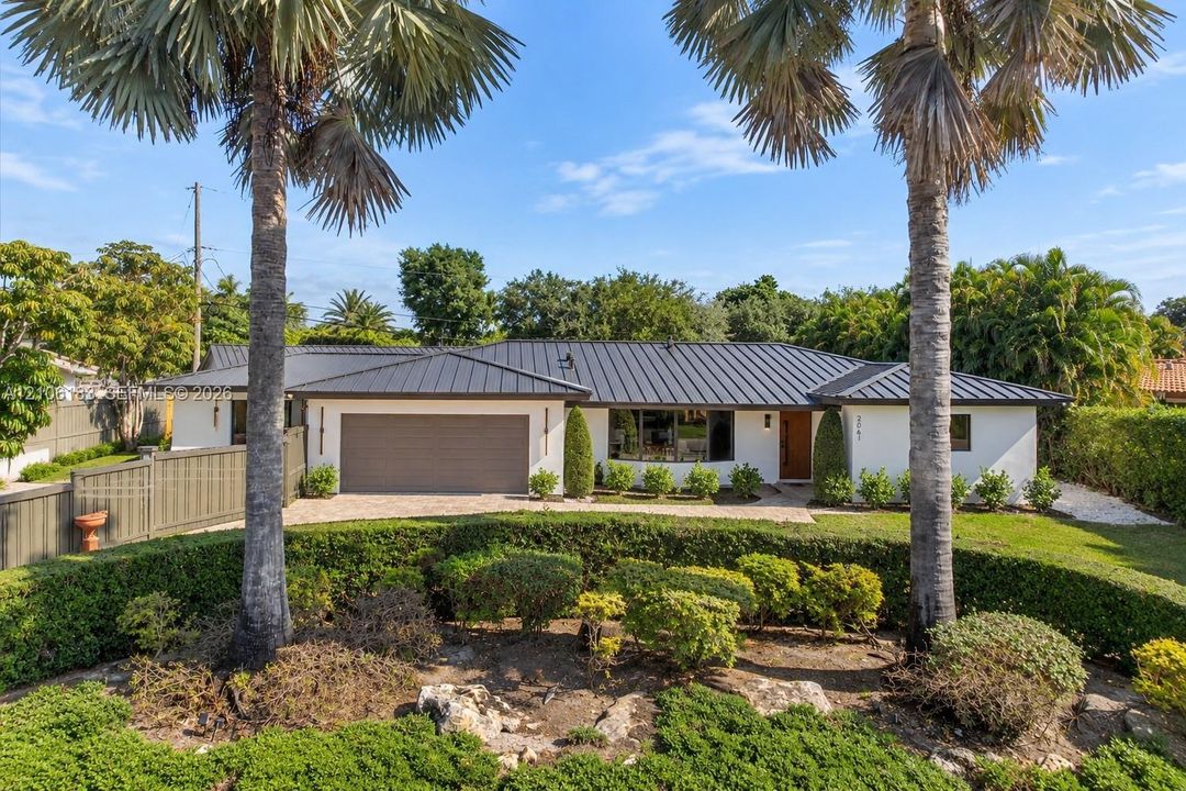 Single-story residence featuring a standing-seam metal roof, stucco exterior, attached garage, stone-paved driveway, and mature landscaping