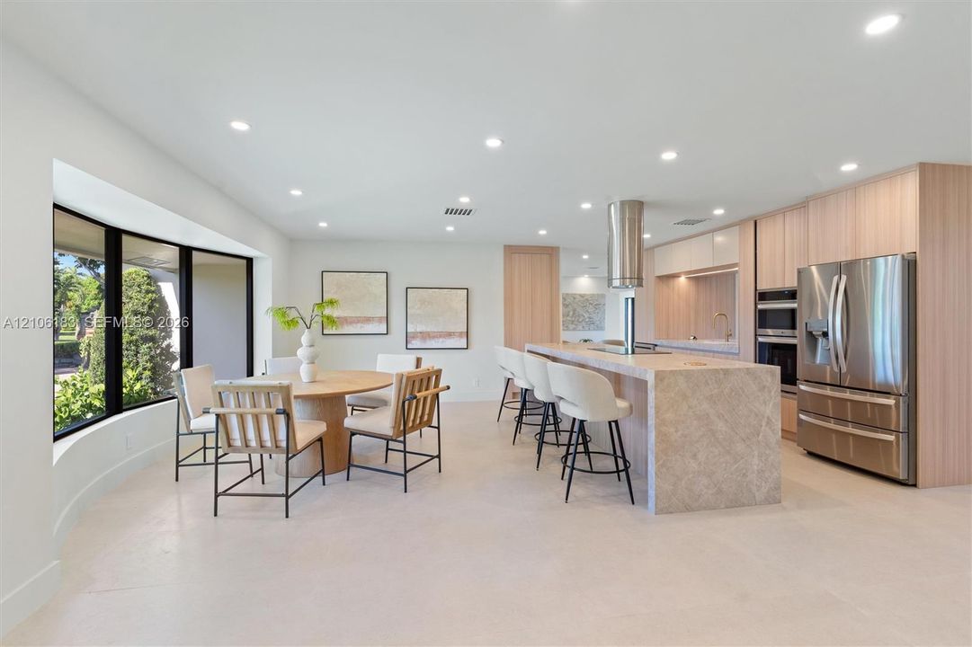Modern kitchen featuring light wood-finish cabinetry, a large stone-surfaced island with a cylindrical exhaust hood, stainless steel appliances, recessed ceiling lighting, and expansive floor-to-ceiling windows