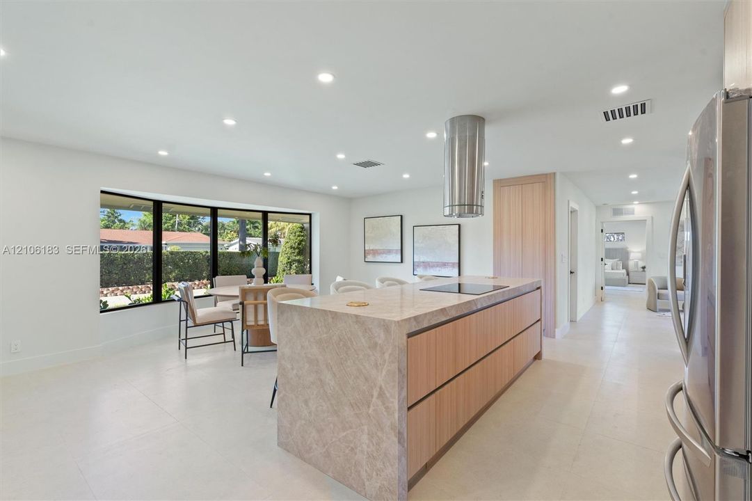 Kitchen featuring a waterfall-edge center island, stone countertops, flat-panel light wood cabinetry, a cylindrical stainless steel range hood, and large windows with black frames