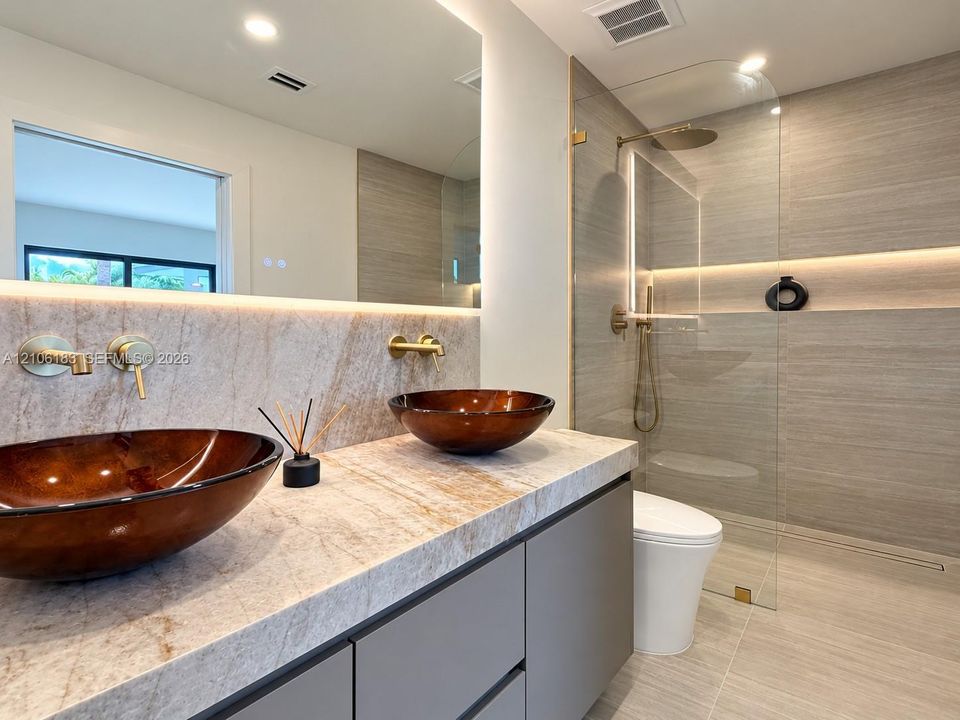 Bathroom vanity featuring a stone countertop, dual glass vessel sinks, wall-mounted gold-tone faucets, and a walk-in shower with floor-to-ceiling tile and integrated lighting