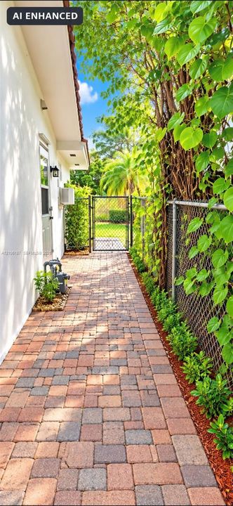 Side yard featuring a multi-tone paver walkway, metal chain-link fencing, a metal gate, and exterior wall-mounted lighting