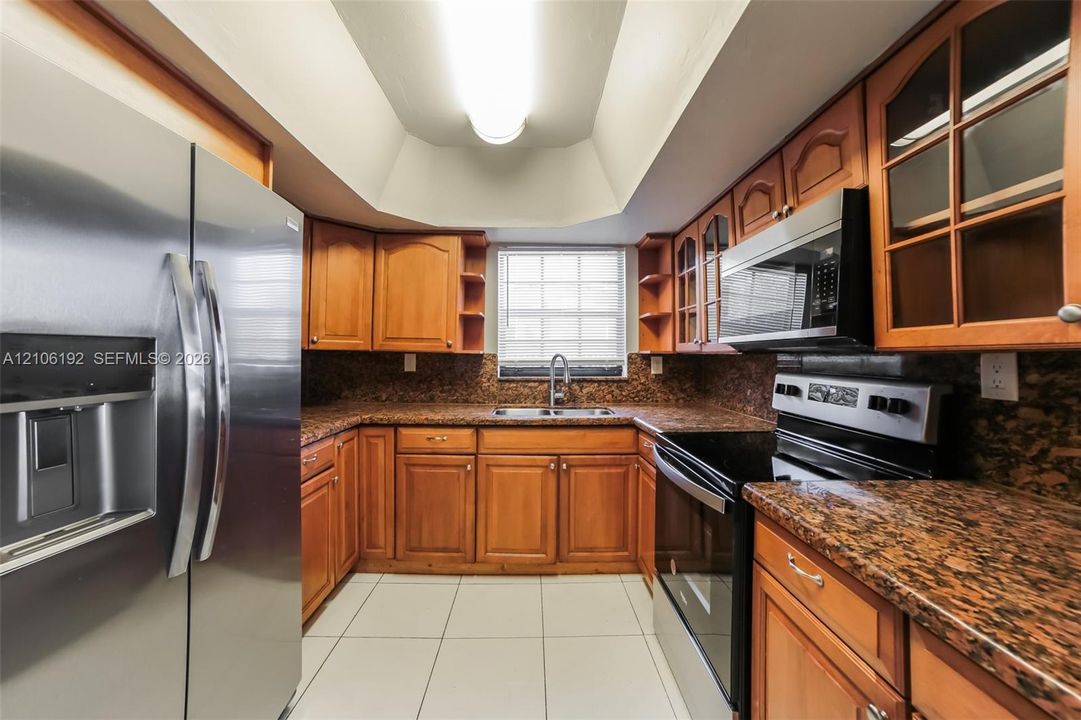 Kitchen featuring warm-tone cabinetry, stone-finish countertops, stainless steel appliances, tile flooring, and a tray ceiling with recessed lighting