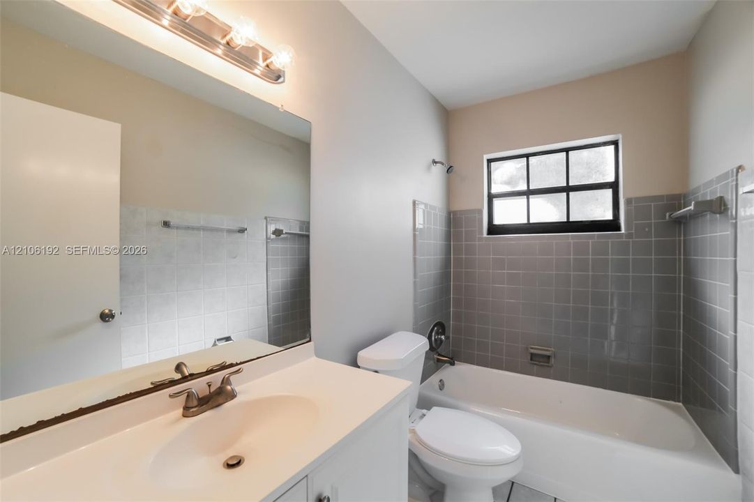 Bathroom featuring a vanity with integrated sink, expansive wall-mounted mirror, multi-bulb light fixture, and a tub-shower combination with gray tile surround and window