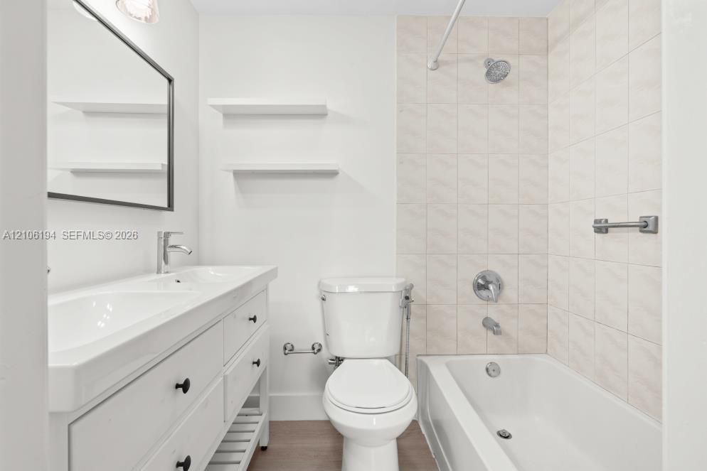 Bathroom featuring a dual-sink vanity with matte black hardware, wall-mounted open shelving, a bathtub and shower combination with light-toned wall tile, and wood-finish flooring