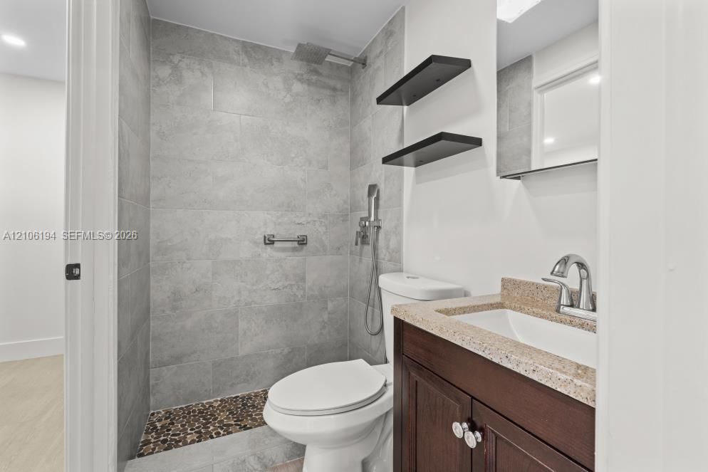 Walk-in shower featuring gray stone-finish tile surround, integrated pebble tile floor, and rainfall showerhead, complemented by a dark wood-finish vanity with a stone-finish countertop, dual-handle faucet, and built-in wall shelving