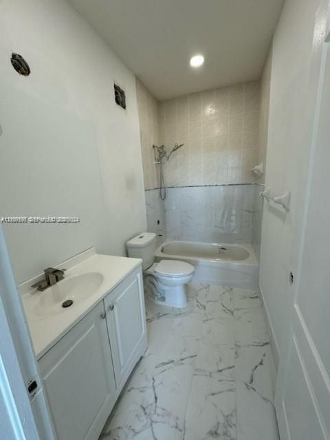 Bathroom featuring a vanity with an integrated sink, polished faucet, white shaker-style cabinetry, and a combination tub and shower with light-toned tiled surrounds and veined stone-patterned floor tile
