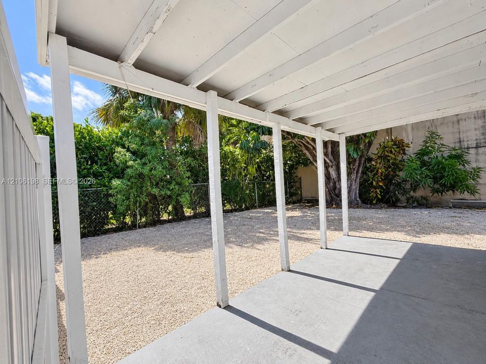 Covered concrete patio featuring a white support structure, gravel yard, and perimeter chain-link fencing