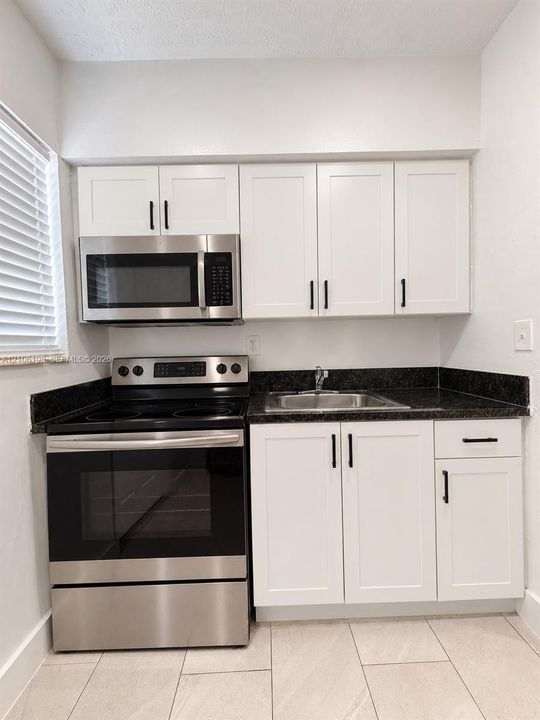 Kitchen area featuring white shaker-style cabinetry, dark stone-finish countertops, stainless steel appliances, and large-format neutral floor tile