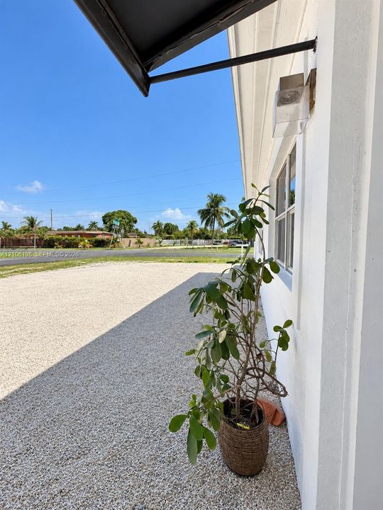 Exterior wall featuring white siding, a fixed-pane window with a frame, an attached metal awning, and an exterior wall-mounted light fixture, complemented by a gravel surface