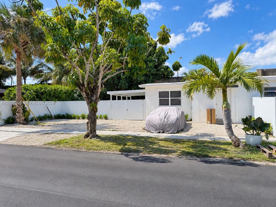 White exterior facade featuring a covered carport, gravel front yard, white perimeter fencing, and mature landscaping