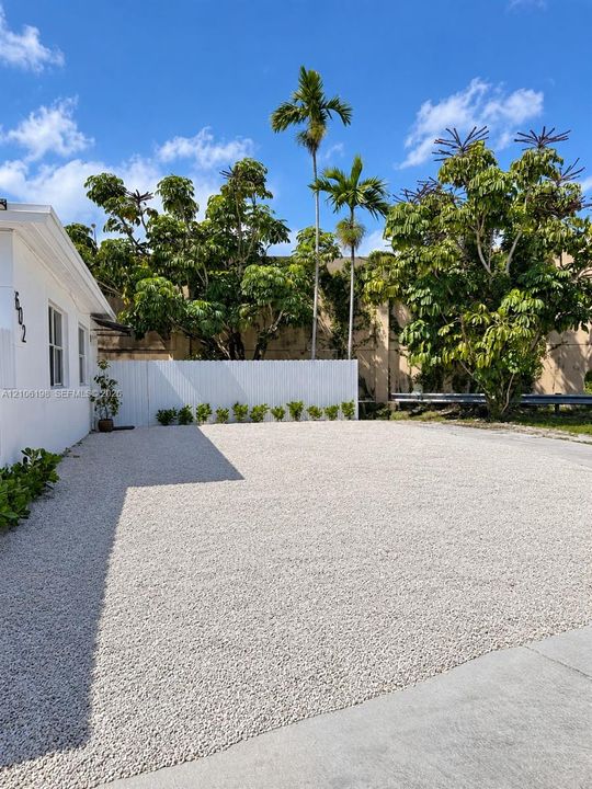 Expansive gravel parking area featuring a white privacy fence, low-maintenance landscaping, and mature palm trees