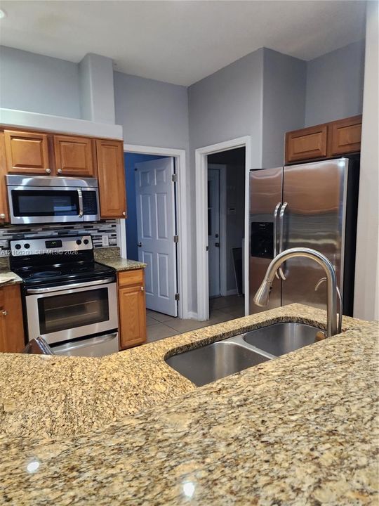 Kitchen featuring stone countertops, stainless steel appliances, wood cabinetry, a dual-basin undermount sink with a gooseneck faucet, and a multi-toned mosaic tile backsplash