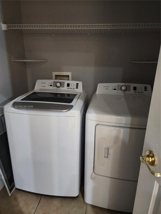 Laundry area featuring white top-load washer and dryer, neutral-tone tile flooring, and overhead wire shelving