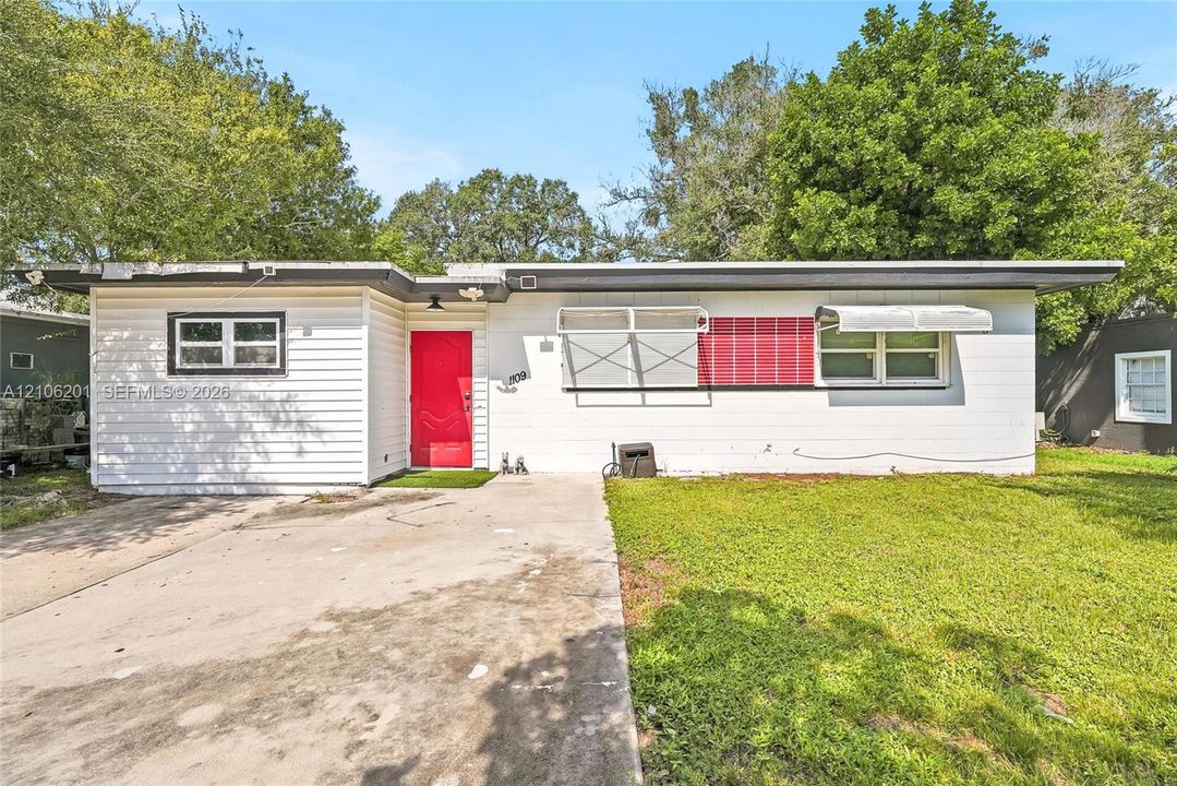 Single-story residence featuring horizontal siding, a bold red entry door, metal window awnings, and a concrete driveway