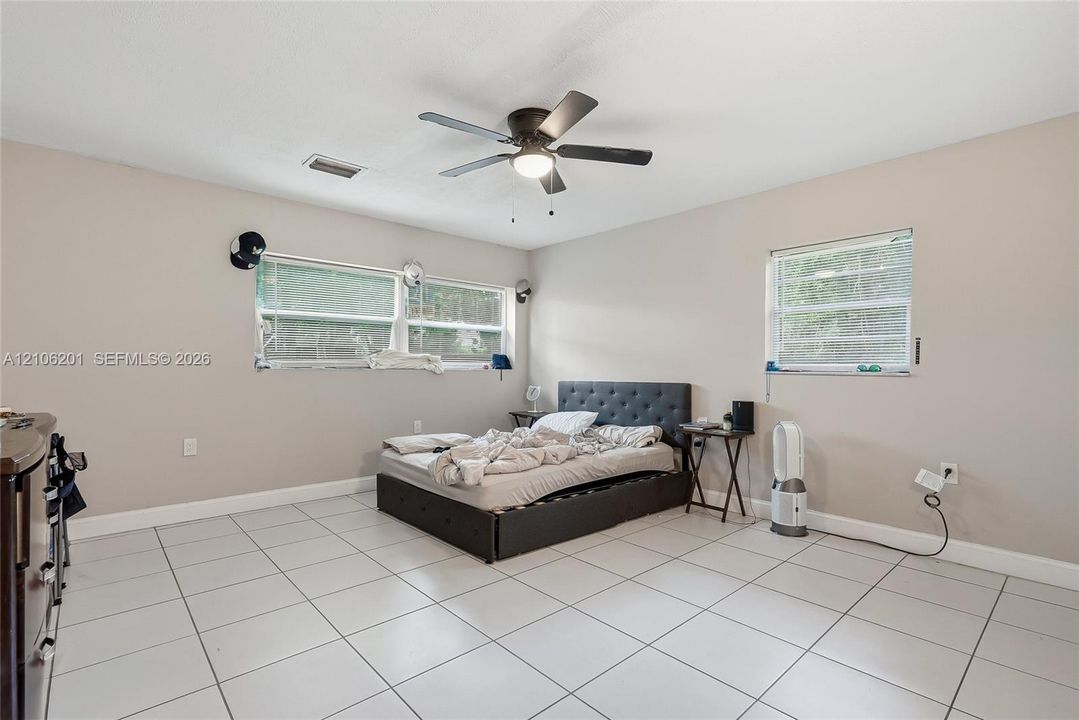 Neutral-toned interior featuring large-format ceramic floor tiles, three windows with horizontal blinds, and a central ceiling fan with an integrated light fixture