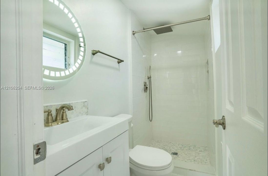 Bathroom featuring a white vanity cabinet, illuminated circular mirror, shower with white tiled surround, rain-style showerhead, and pebble-finish shower floor