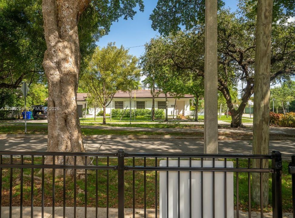 Single-story residence featuring a light-toned exterior, dark-toned hip roof, and mature trees situated on a parcel with metal perimeter fencing