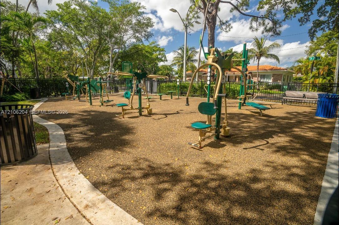 Outdoor fitness area featuring a rubberized surface, multiple static exercise stations, bench seating, and perimeter fencing shaded by mature trees