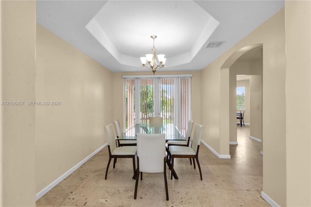 Tray ceiling with central chandelier, neutral wall tones, large-format stone-finish flooring, and glass doors with vertical blinds