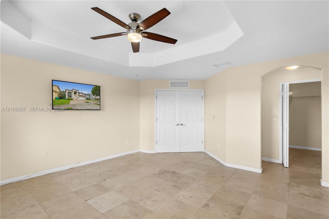 Tray ceiling featuring a central five-blade ceiling fan with integrated lighting, tile flooring, double-panel doors, and an arched entry to a walk-in closet