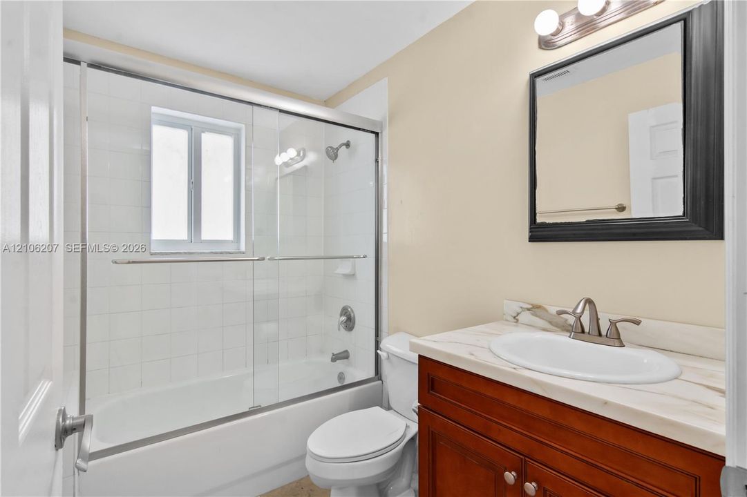 Bathroom featuring a wood-finish vanity with a stone-top integrated sink, a glass-enclosed shower and tub combination with tiled surround, and a wall-mounted framed mirror