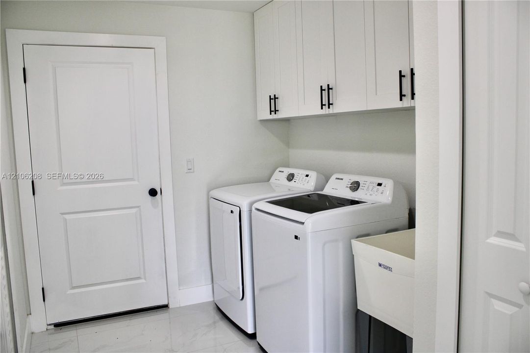 Laundry room featuring white shaker-style wall cabinets with matte black hardware, a utility sink, and large-format tile flooring