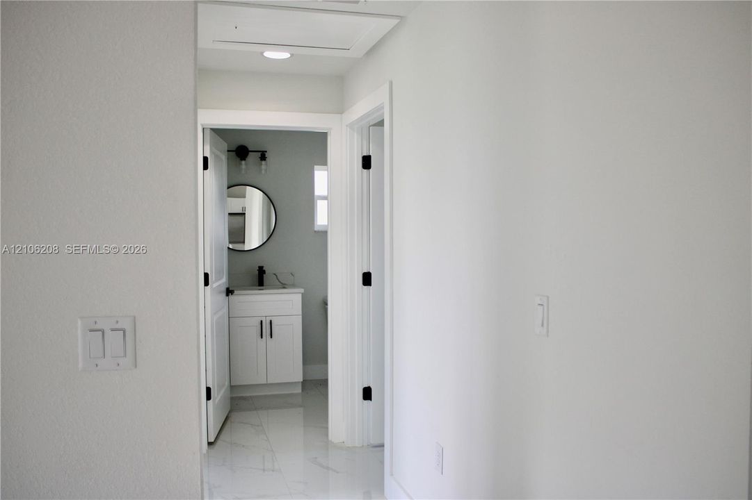 Hallway featuring light-colored tile flooring and recessed ceiling lighting, revealing a bathroom with a white vanity, matte black hardware, and a circular mirror