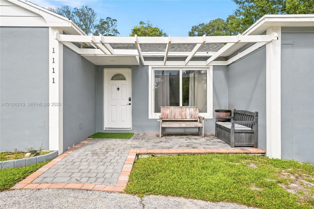 Exterior entryway featuring a white pergola, paver patio, brick edging, and a single-light panel front door