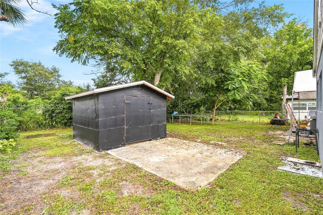 Backyard area featuring a structural storage shed, concrete foundation pad, chain-link fencing, and mature trees