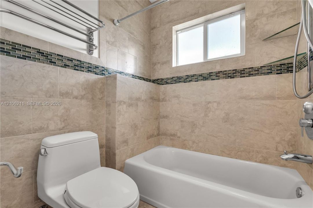 Full-height wall tiling featuring natural stone-finish tiles and a decorative glass mosaic accent band, white bathtub with chrome plumbing fixtures, integrated glass shelving, and a wall-mounted metal towel rack