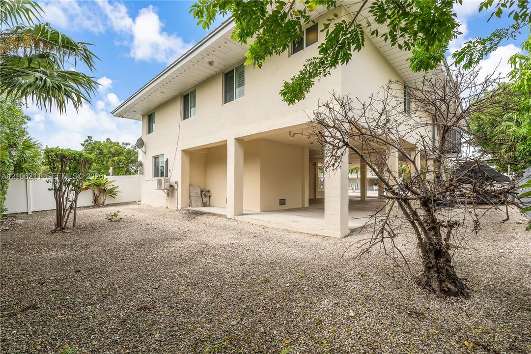 Two-story exterior featuring a cream-colored stucco facade, raised construction with open-air lower level, gravel grounds, and perimeter privacy fencing