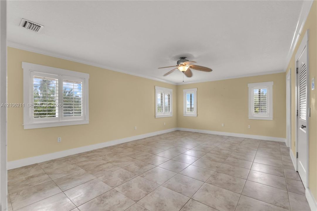 Neutral-toned interior room featuring large-format ceramic tile flooring, plantation shutters, and a ceiling fan with integrated lighting