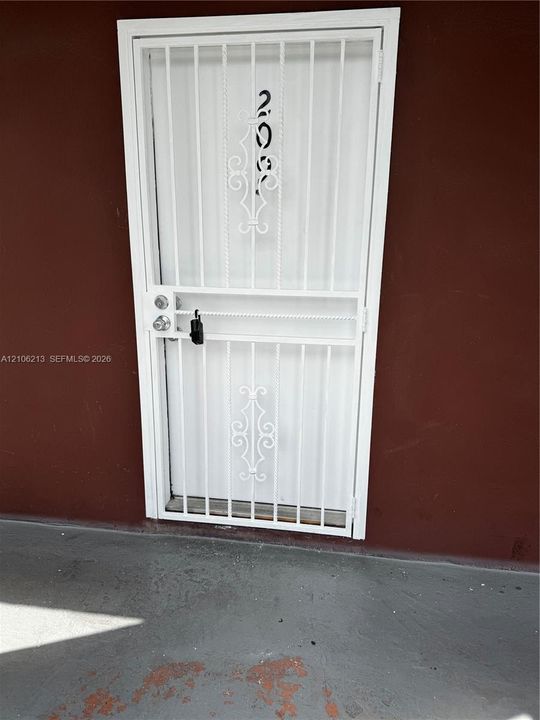 White metal security screen door featuring decorative scrollwork, a vertical bar pattern, and a dual-locking mechanism
