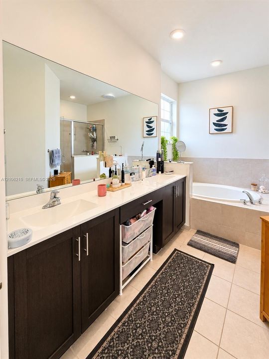 Bathroom featuring a dual-sink vanity with dark cabinetry, stone-finish countertops, full-width mirror, tile flooring, and a deep soaking tub
