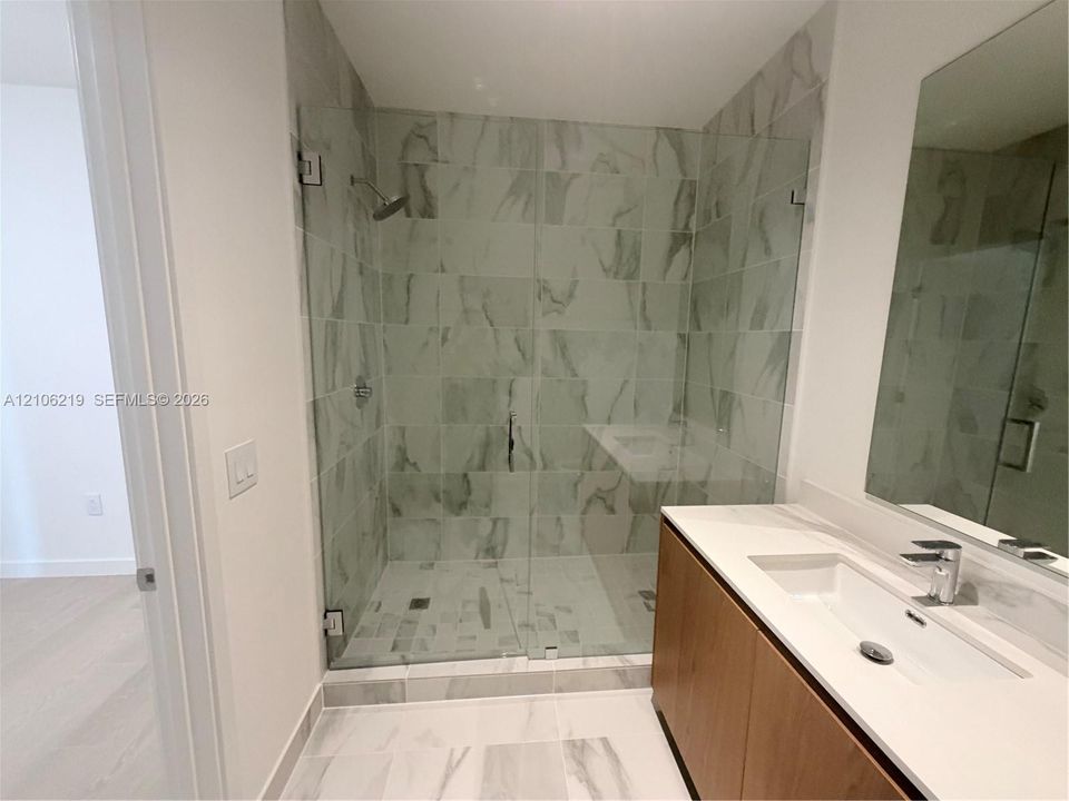 Walk-in shower featuring large-format tile surround, glass enclosure, and rainfall showerhead, paired with a vanity offering a rectangular integrated sink, white countertop, and wood-finish cabinetry
