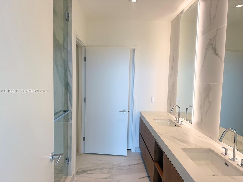 Dual-sink vanity with wood-finish cabinetry, stone-pattern countertop, integrated vertical LED mirror lighting, and walk-in glass shower enclosure with floor-to-ceiling stone-pattern tile surround