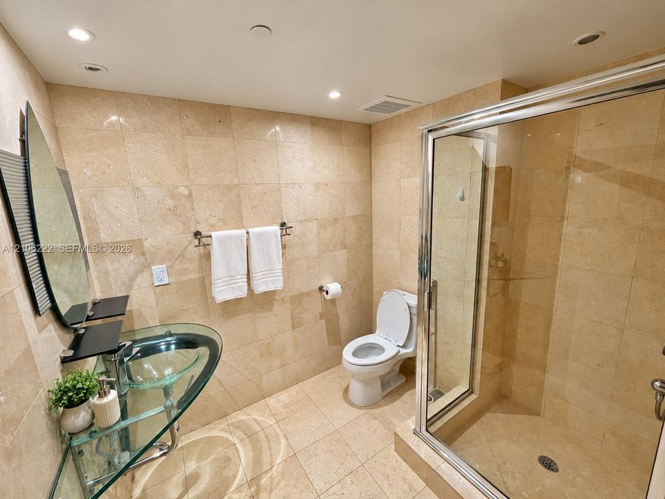 Full-height stone tile wall finishes with matching floor tiling, glass pedestal sink, frameless glass-enclosed shower, and recessed ceiling lighting