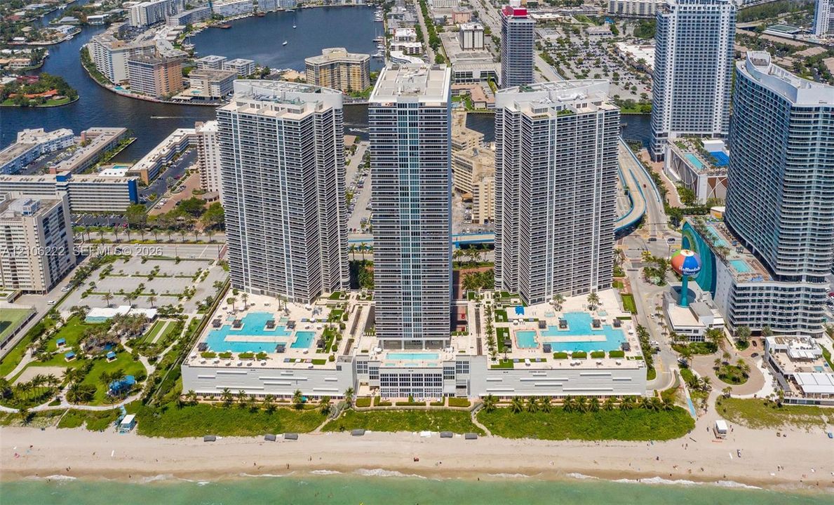 Aerial perspective of oceanfront high-rise residential complex featuring multiple swimming pools, spacious sun decks, extensive podium-level hardscaping, and direct beach access