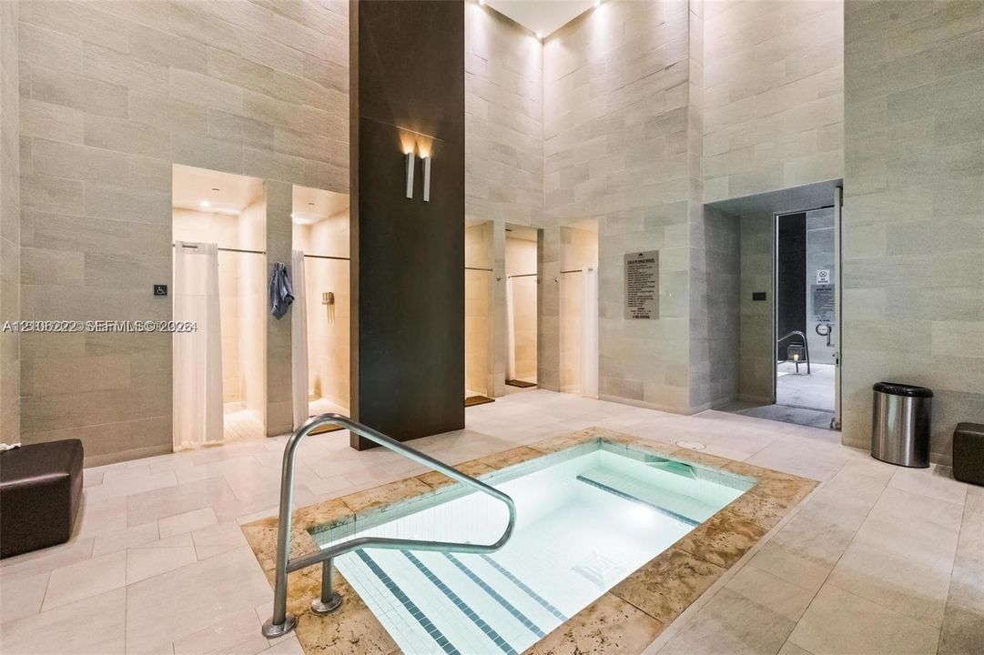 Soaking pool featuring a stone tile surround and metal grab rail, floor-to-ceiling stone wall cladding, and multiple private shower enclosures