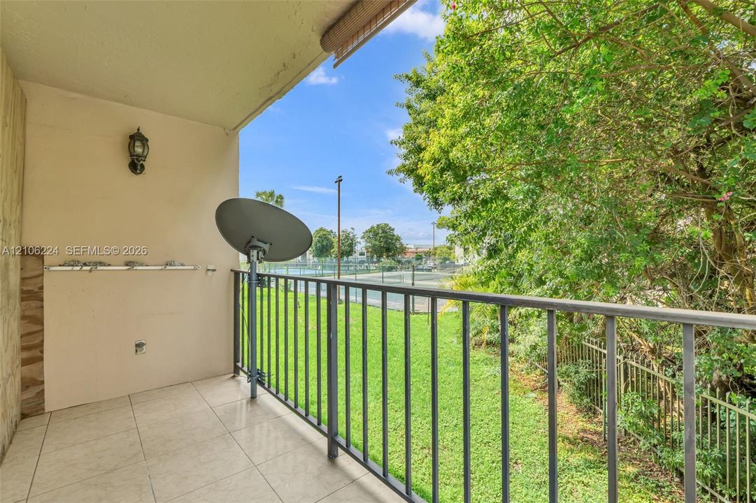 Covered balcony featuring tile flooring, metal railing, wall-mounted exterior light fixture, and mature tree backdrop