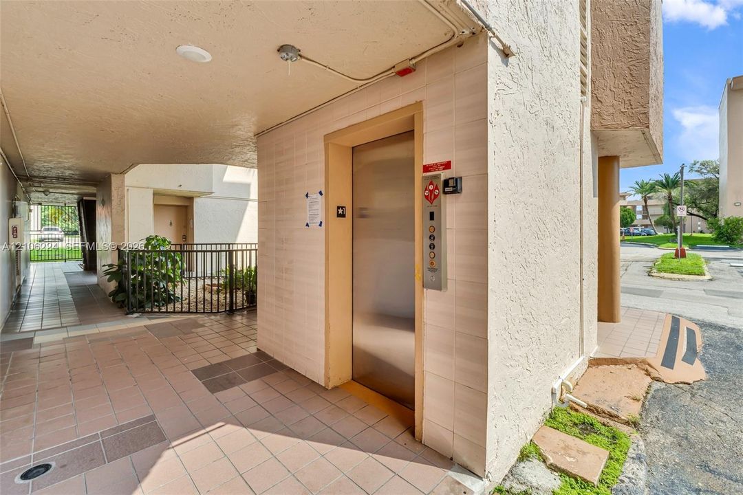 Building exterior featuring a tiled ground-level corridor, metallic elevator doors, and a wrought iron perimeter fence