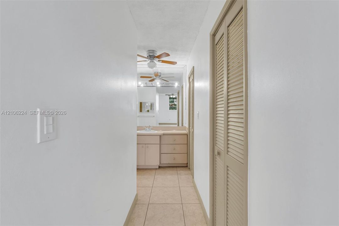 Hallway featuring tile flooring, dual louvered closet doors, and a vanity with integrated cabinetry, capped by a large wall-mounted mirror and ceiling-mounted paddle fan