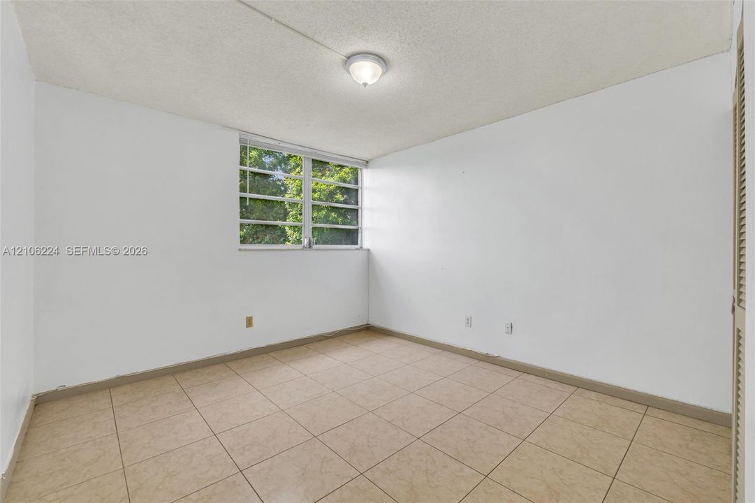 Neutral-toned tiled flooring, textured ceiling with flush-mount lighting, multi-pane window, and louvered closet doors